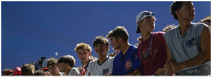 A group of people wearing casual clothing stand closely together outdoors under a clear evening sky. Description generated by AI