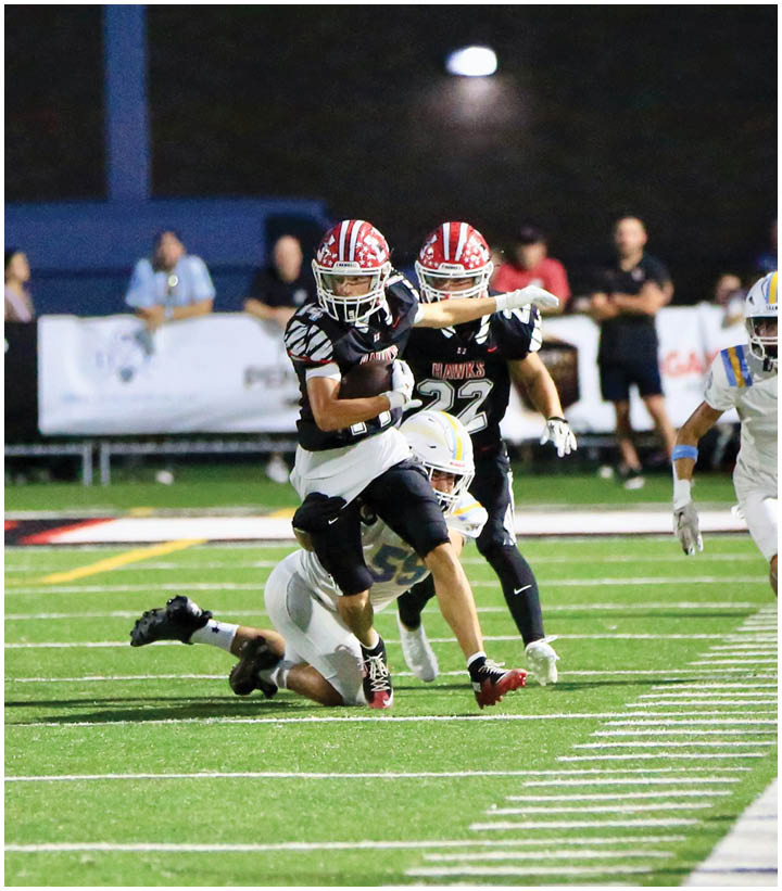 Two football players in black uniforms with red helmets are running on a field while a player in a white uniform attempts to tackle one of them. Description generated by AI