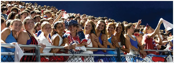 A large group of people wearing patriotic clothing and accessories are gathered closely together in stadium bleachers under a dark sky, smiling and posing for the camera. Description generated by AI