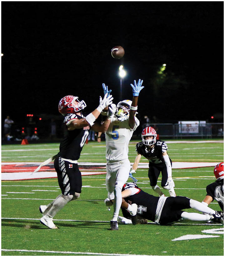 Two people in football uniforms jump with arms extended to catch a football while others on the field watch or lie on the ground during a nighttime game. Description generated by AI