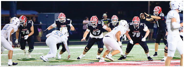 People in football uniforms are positioned on a field preparing for a play during a game at night. Description generated by AI
