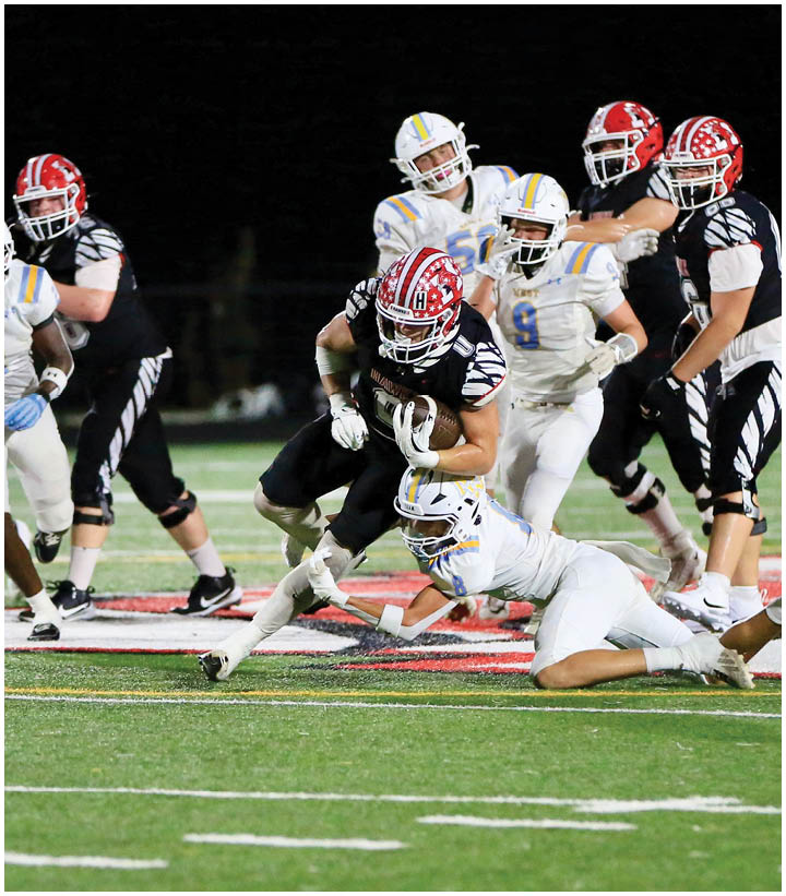 A group of people in football uniforms are playing on a field at night with one person in black running with the ball while others in white attempt to tackle or block. Description generated by AI