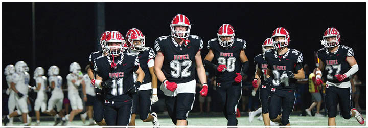 A group of people wearing black and red football uniforms with helmets are walking together on a field at night with another group in white uniforms in the background. Description generated by AI