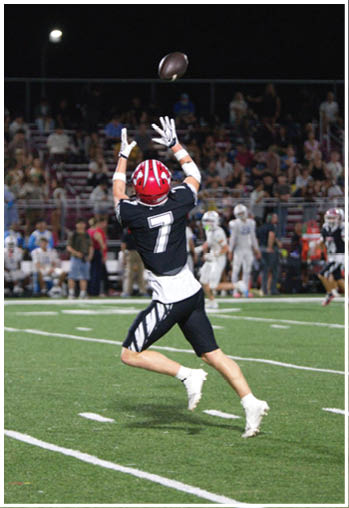 A person wearing a black and white football uniform with the number seven is reaching up to catch a football on a lit field with a crowd in the background. Description generated by AI