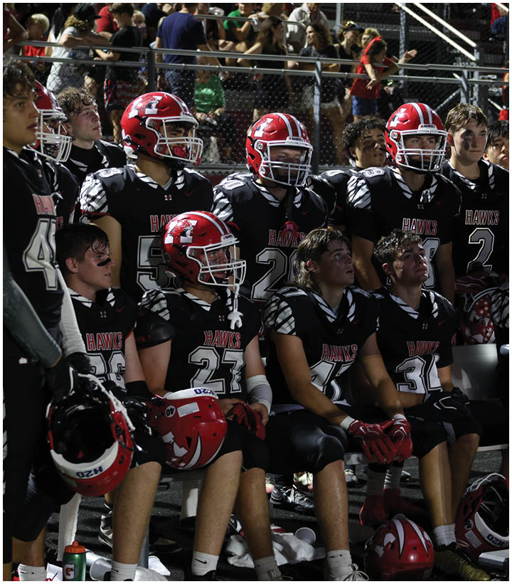 A group of people wearing black and red football uniforms with helmets are sitting and standing on the sidelines of a sports field at night. Description generated by AI