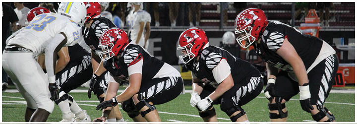 People in football uniforms and helmets are lined up on a field in a crouched position preparing for a play. Description generated by AI