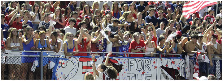 A group of people in athletic clothing stands behind a fence holding a large decorated banner with “25 FOR THE" written on it while others in front of the fence appear to be interacting with them. Description generated by AI