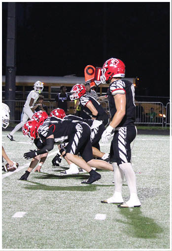 People on a football field at night include a player in a black and white uniform with a red helmet and two others standing near a metal fence in the background. Description generated by AI