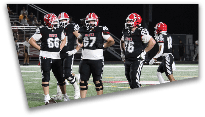 Four people in black and white football uniforms with red helmets stand on a field with one person holding a football on the ground. Description generated by AI