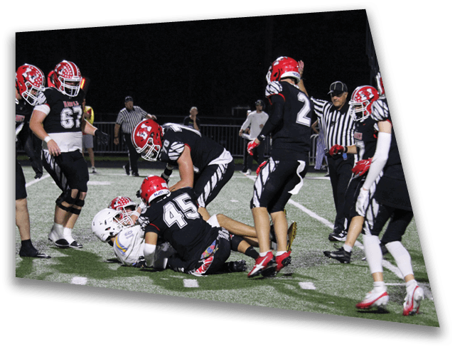 A group of people wearing black and red football uniforms are on a field at night with one person in white being tackled to the ground. Description generated by AI