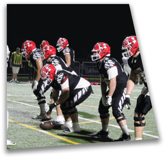 Two people in football uniforms with red helmets and black jerseys are positioned on a football field during a game with spectators and other players in the background. Description generated by AI