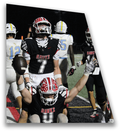 Several football players in black and white uniforms with “HAWKS" on the jerseys are on a field at night, with one player raising an arm and another player in a white and blue uniform visible in the background. Description generated by AI