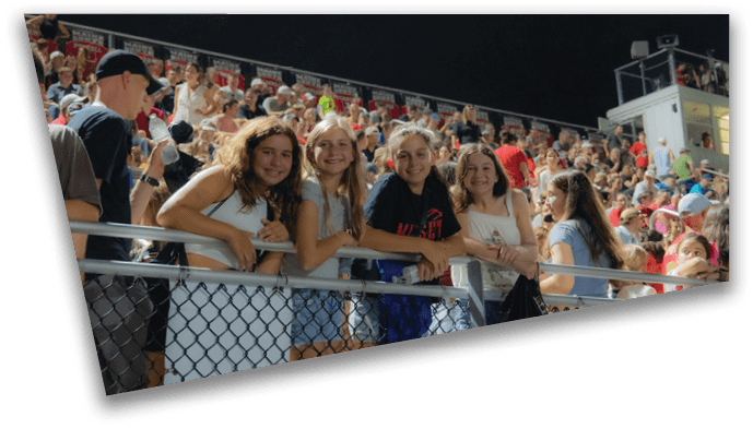 Four smiling people leaning on a railing in front of a large crowd at a nighttime outdoor event. Description generated by AI