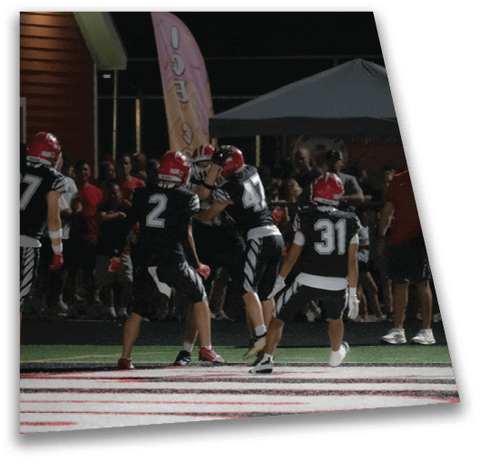 People wearing black sports uniforms and red helmets are playing football on a field at night with a crowd watching in the background. Description generated by AI
