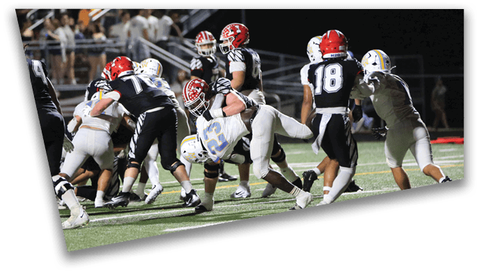 People wearing football uniforms are engaged in a tackle during a game on a grassy field under stadium lights. Description generated by AI