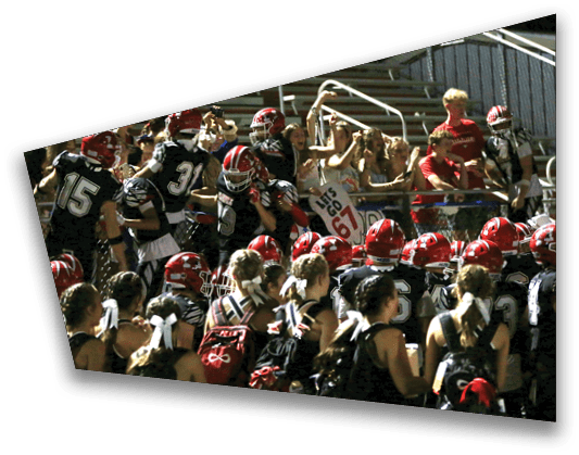 A group of people wearing sports uniforms and helmets are gathered closely together near bleachers with some people in casual clothing standing behind them. Description generated by AI