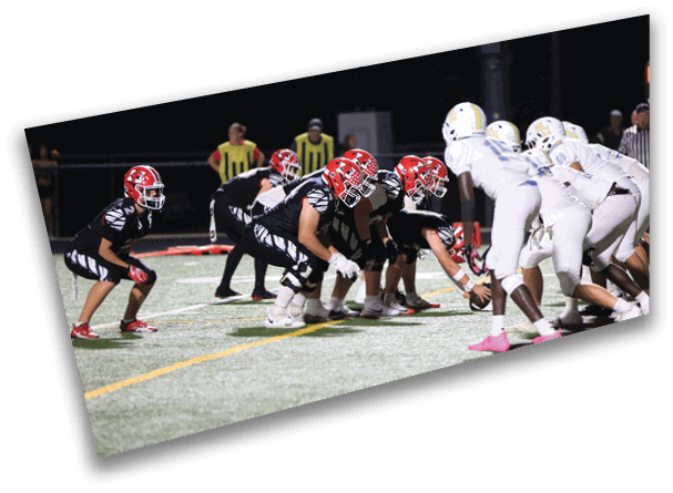 Two opposing American football teams in black and white uniforms are lined up at the line of scrimmage on a field under stadium lights with referees and officials in the background. Description generated by AI