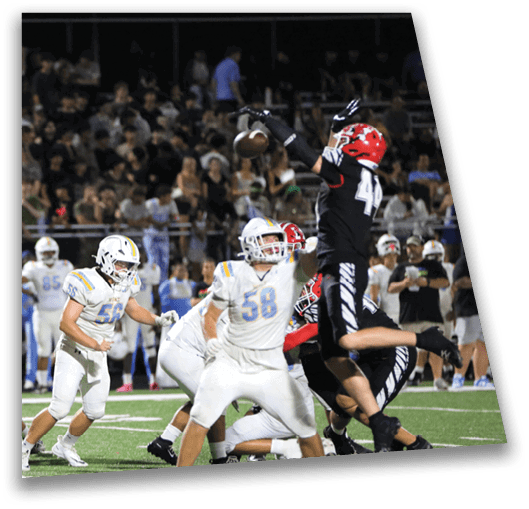 A group of people in football uniforms are actively playing a game on a field with a crowd watching in the background. Description generated by AI