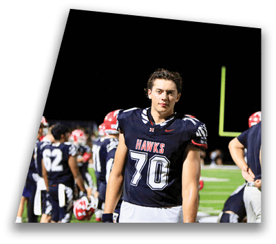 A person wearing a dark football uniform with the word “HAWKS" and the number 70 stands on a field at night with other players in the background. Description generated by AI