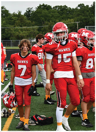 Three people wearing red football helmets and red jerseys stand outdoors with trees in the background. Description generated by AI