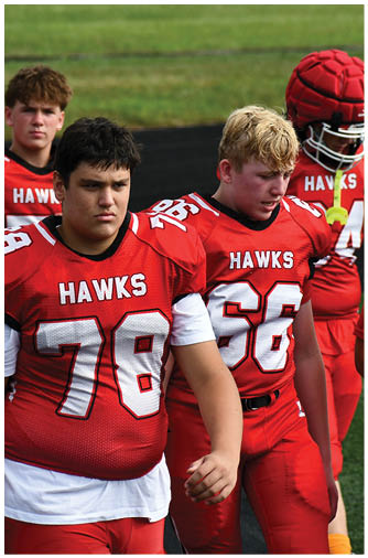 People wearing red football uniforms with numbers and orange socks are standing outdoors on a paved surface. Description generated by AI