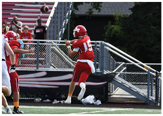 A person wearing a red football uniform and helmet is preparing to catch a football near metal bleachers and a fence with trees in the background. Description generated by AI