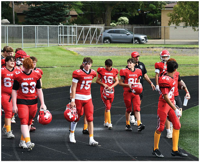 A group of people wearing red football uniforms with “HAWKS" on the jerseys are standing and walking on a track near a grassy field with a car and fence in the background. Description generated by AI