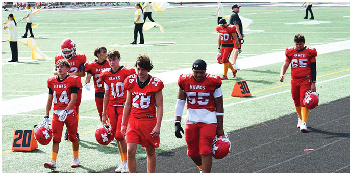 A group of people wearing red football uniforms walk together on a field near yard markers. Description generated by AI