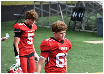 A person wearing a red football jersey with the number 61 and the word “HAWKS" stands on a green field near empty bleachers and chairs. Description generated by AI
