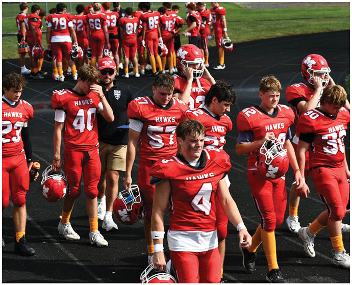 A group of people wearing red football uniforms with “HAWKS" on the jerseys are standing on a track, some holding helmets and others with helmets on. Description generated by AI