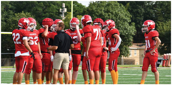 A group of people wearing red football uniforms and helmets are gathered on a green field with one person in black shirt and khaki shorts standing among them. Description generated by AI