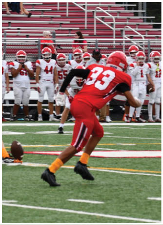 A person wearing red sports gear and orange socks is running on a green football field with white and yellow lines, while a group of people stand in the background. Description generated by AI
