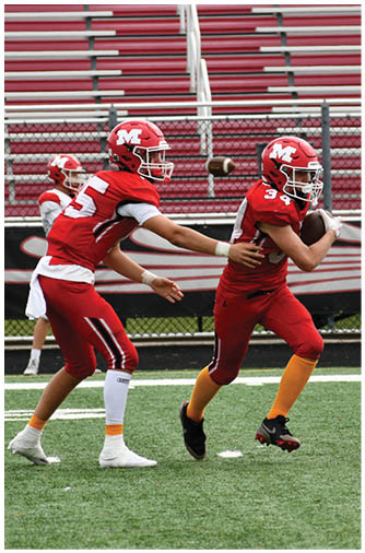 A person wearing a red football uniform and helmet is poised to throw a football on a field with empty bleachers in the background. Description generated by AI