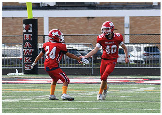 Two people in red football uniforms with helmets are on a field, with one reaching out to the other near a goalpost and a scoreboard in the background. Description generated by AI