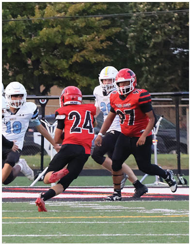 Three people in football uniforms are on a field with one person in a red jersey standing near a fence and two others in white jerseys running on the turf. Description generated by AI