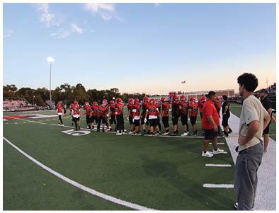 A group of people wearing red and black football uniforms are lined up on a football field under a clear sky during sunset. Description generated by AI