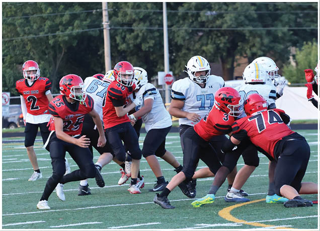 People wearing football helmets and uniforms are actively engaged in a game on a field with trees and a stop sign visible in the background. Description generated by AI