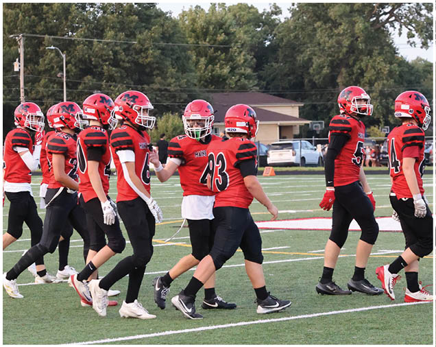 A group of people wearing red and black football uniforms and helmets are standing together on a football field with trees and buildings in the background. Description generated by AI