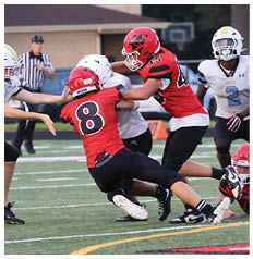 Four people in football uniforms are engaged in a play on a field with one person in a red jersey tackling another in a white jersey while a referee watches in the background. Description generated by AI