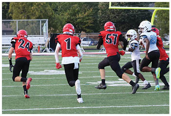 Three people in red football uniforms with helmets are running on a green football field with white yard lines and a goalpost in the background. Description generated by AI
