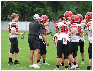 An adult wearing a cap is holding a football and interacting with a group of children in football uniforms and helmets on a grassy field. Description generated by AI