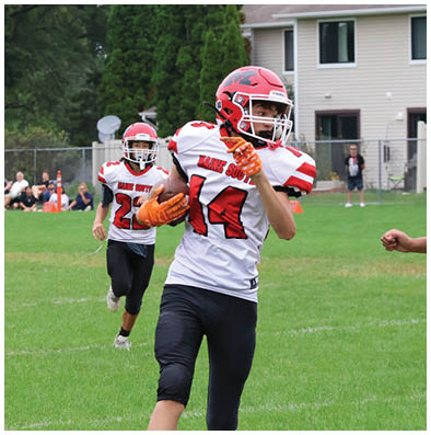 Two people wearing red helmets and white sports jerseys are playing football on a grassy field with spectators and a house in the background. Description generated by AI