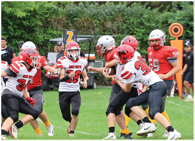 Several people wearing football helmets and uniforms are engaged in a play on a grassy field with a chain marker visible in the background. Description generated by AI