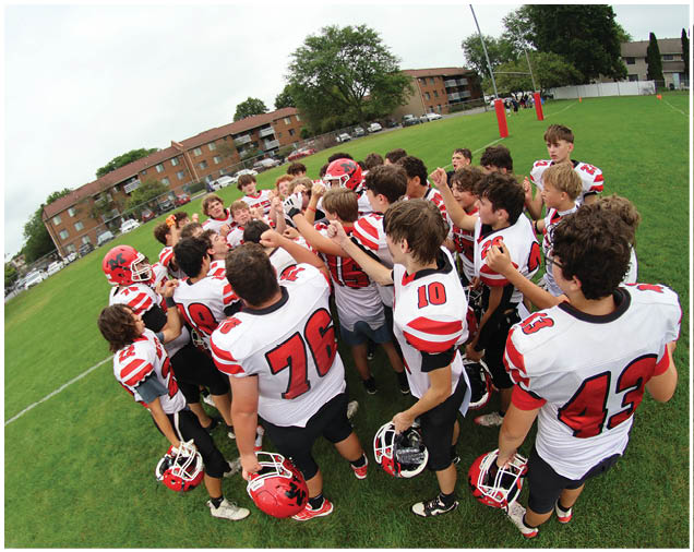 A group of people wearing red and white football uniforms are gathered closely together on a grassy field near residential buildings. Description generated by AI