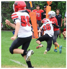 Two people in football gear are engaged in a tackle on a grassy field while others, including a referee and spectators, watch in the background. Description generated by AI