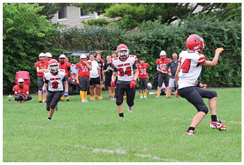 People wearing red and white football uniforms are playing on a grassy field with trees and houses in the background. Description generated by AI