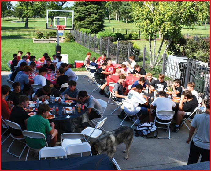 A large group of people sitting at multiple outdoor tables on a basketball court enjoying a meal together with a dog nearby. Description generated by AI