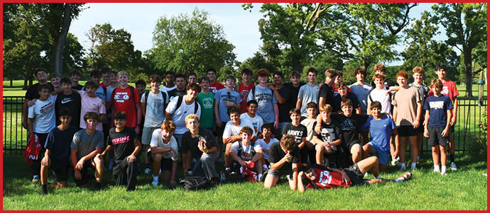 A large group of people wearing casual athletic clothing is gathered outdoors on grass in front of a fence with trees in the background. Description generated by AI