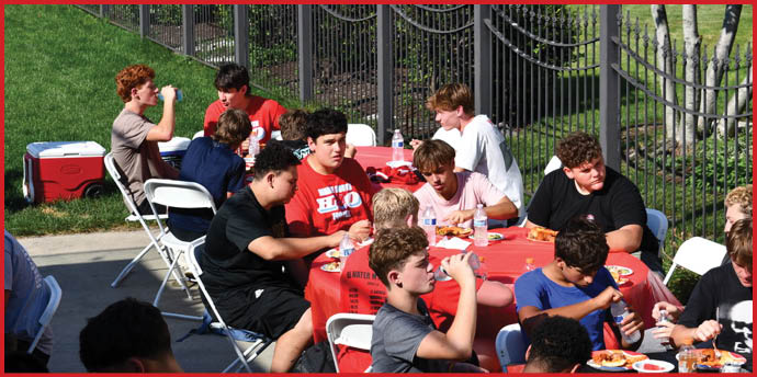 A group of people sitting around a red tablecloth-covered table outdoors, eating and drinking while engaging in conversation. Description generated by AI