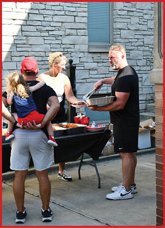 Three people stand close together in front of a stone wall, with one person wearing a red cap and holding a child with long hair, while another person with light hair and sunglasses on their head looks to the side. Description generated by AI